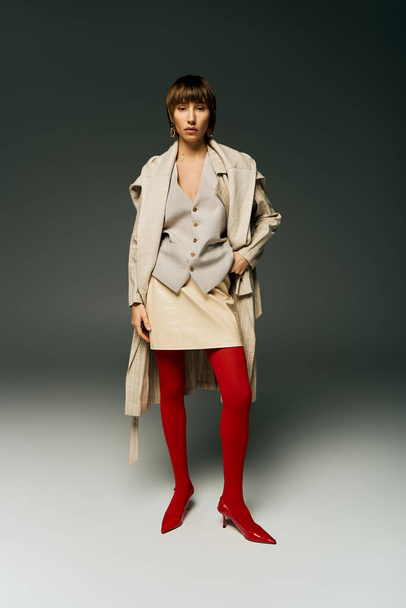 Uma jovem com cabelo curto posa em um casaco de trincheira e meia-calça vermelha em um estúdio. - Foto, Imagem