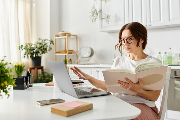 Middelbare leeftijd vrouw verdiept in een boek, multitasking met een laptop. - Foto, afbeelding