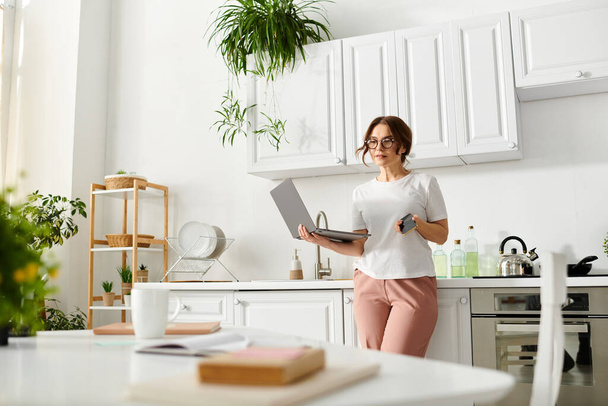 A middle-aged woman stands in the kitchen, holding a laptop. - Photo, Image