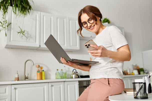 Femme d'âge moyen assis sur le comptoir, dactylographiant sur ordinateur portable. - Photo, image