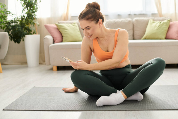 Kobieta w średnim wieku siedzi na macie do jogi, pochłonięta telefonem. - Zdjęcie, obraz