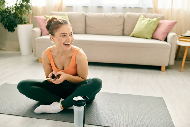 Middelbare leeftijd vrouw zit op yoga mat, met een mobiele telefoon. - Foto, afbeelding