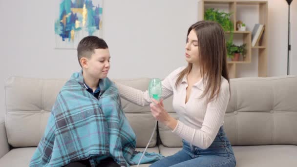 Äiti auttaa sairasta poikaa käyttämään sumutinta syleillessään häntä kotona sohvalla. Nainen tekee hengitys laitteiden kanssa pojalle - Materiaali, video