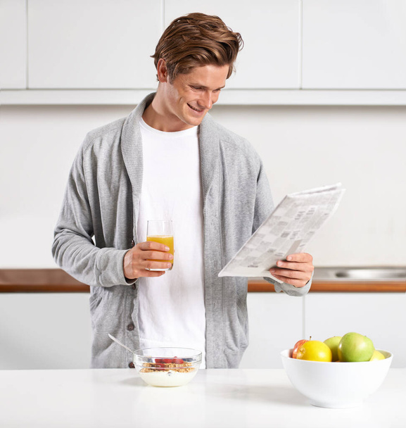 Breakfast, juice and man with newspaper in kitchen for information at modern apartment. Nutrition, cereal and young male person drinking healthy beverage and reading public journalism at home - Photo, Image