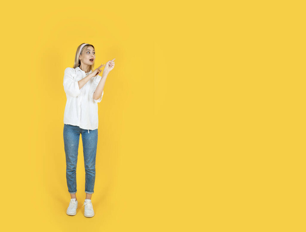 Positive happy 20s blonde woman demonstrating pointing copy space. Full length body size attractive cheerful girl looking aside, isolated yellow studio background. Advertisement concept idea photo. - Photo, Image
