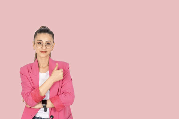 Бізнес-леді показує великий палець, портрет молодої кавказької брюнетки бізнес-леді показує великий палець вгору. Носіння окулярів і рожевий офіційний піджак. Ізольований рожевий фон студії, простір для копіювання. Дивлячись камеру - Фото, зображення
