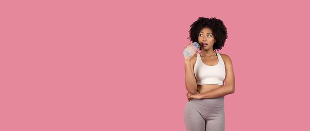 Mulher negra jovem focada na aptidão com cabelo encaracolado, vestindo roupas esportivas, segurando garrafa de água, olhando para o espaço livre pensativamente contra o fundo rosa, panorama - Foto, Imagem