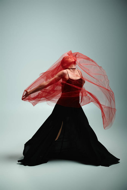 Een jonge, mooie ballerina in een zwarte jurk beweegt sierlijk met een vloeiende rode sluier. - Foto, afbeelding