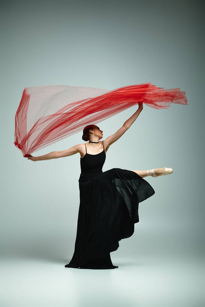 Μια νεαρή μπαλαρίνα με μαύρο φόρεμα κρατά με χάρη ένα ζωηρό κόκκινο μαντήλι.. - Φωτογραφία, εικόνα