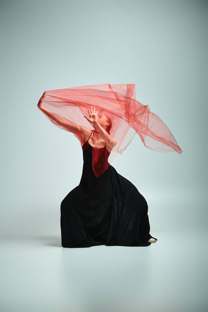 Μια χαριτωμένη μπαλαρίνα με ένα εντυπωσιακό μαύρο φόρεμα κινείται κομψά με ένα κόκκινο πέπλο στο κεφάλι της. - Φωτογραφία, εικόνα