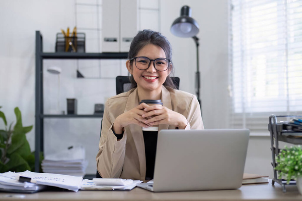 Azjatycka bizneswoman lub księgowa szczęśliwie siedzi i pracuje z laptopem na finansach i administracji biznesowej. Na biurku w biurze ozdobionym zielonymi roślinami.. - Zdjęcie, obraz
