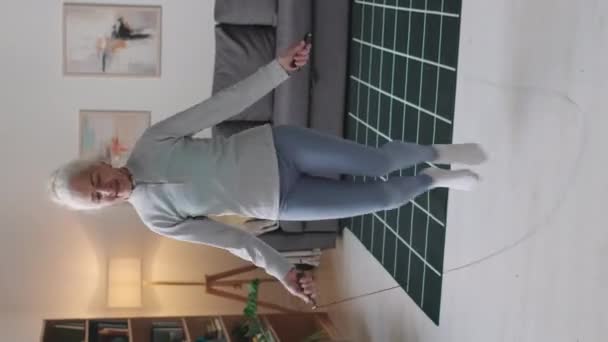 Pionowy pełny strzał aktywnej kaukaskiej seniorki skakanki podczas domowego treningu cardio w nowoczesnym salonie - Materiał filmowy, wideo