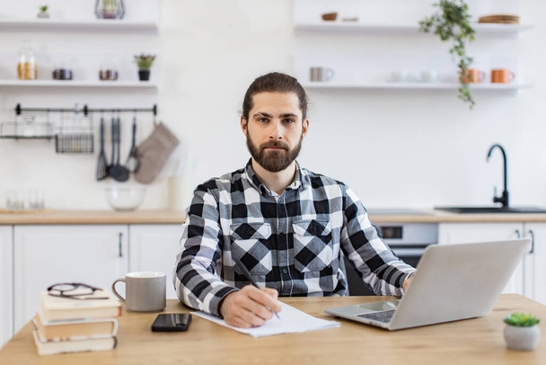 Bearded Caucasian businessman with pen making notes on draft of paper document being placed near laptop on wooden desk. Well-organised man remote employee starting business day in workplace at home. - Foto, Imagem