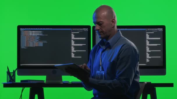 Портрет разработчика программного обеспечения с использованием цифрового планшета и позирование для камеры за столом с мониторами компьютера отображения кода программирования в студии с зеленой стеной хрома ключ - Кадры, видео