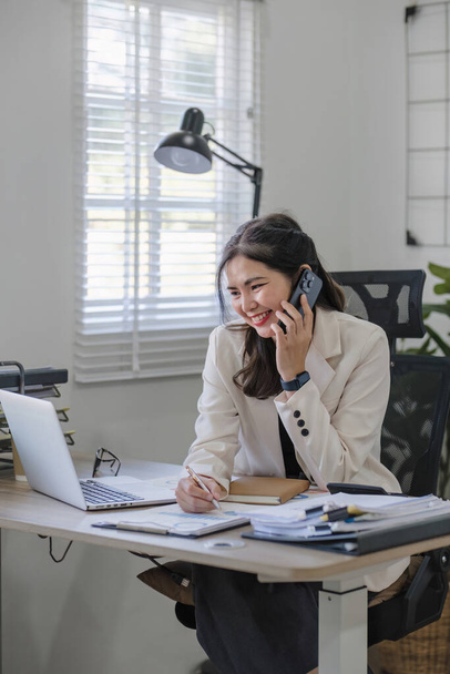 Mladá asijská podnikatelka sedí na telefonu v on-line obchodní jednání pomocí notebooku v moderní domácí kanceláři zdobené stinné zelené rostliny.. - Fotografie, Obrázek