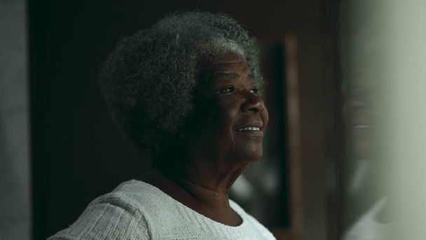 Uma senhora negra idosa contemplativa feliz nos anos 80 olhando pela janela residencial. Candid Thoughtful Senior mulher afro-americana - Foto, Imagem