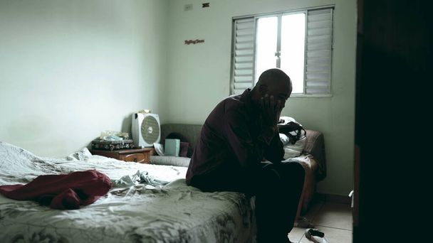 Jeden przygnębiony Murzyn w średnim wieku, siedzący w łóżku, cierpiący na chorobę psychiczną w ponurej sypialni, po pięćdziesiątce Afrykanin zmaga się z ubóstwem, zakrywając twarz. - Zdjęcie, obraz