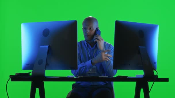 Especialista del centro de datos sentado en el escritorio con monitor de computadora dual y hablando por teléfono móvil contra fondo verde - Imágenes, Vídeo