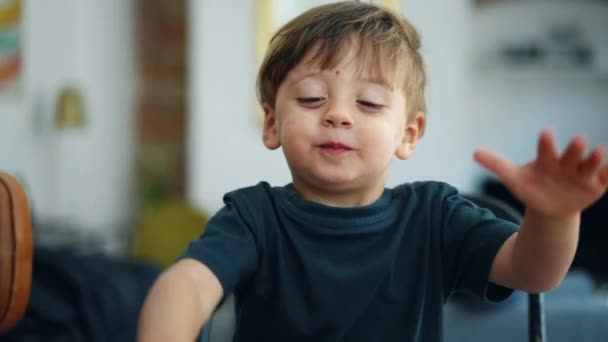 Un niño adorable feliz de dos años sintiendo un momento de alegría genuina cara de cerca sentado en el mostrador de la cocina con vistas a la sala de estar en el fondo. Retrato de niño rubio caucásico - Metraje, vídeo