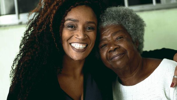 Tender loving moment of African American adult daughter kissing elderly 80s senior mother in the forehead and posing close-up faces smiling at camera - Photo, Image