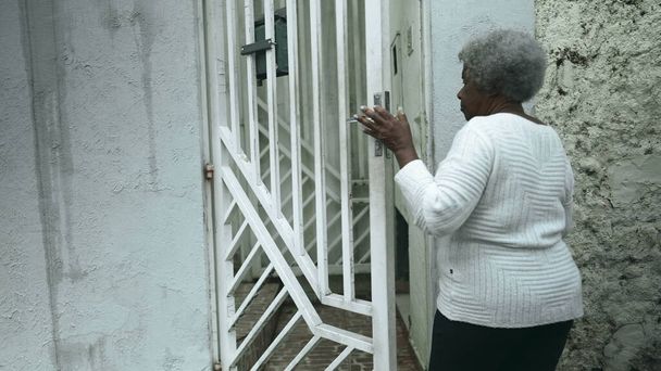 Anciana mujer negra sudamericana llega a casa desde la calle de la acera, abre la puerta principal de la residencia regresando cerrando la puerta detrás de ella, persona mayor de 80 años de ascendencia africana - Foto, imagen