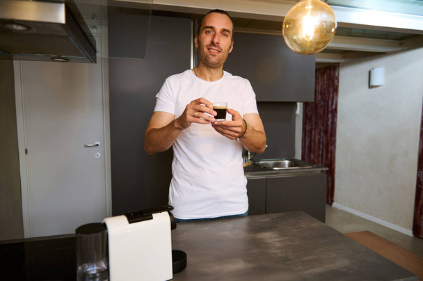 Schöner junger kaukasischer Geschäftsmann in weißem T-Shirt steht am Küchentisch in einem minimalistischen Wohnkücheninterieur und genießt den Duft von Espresso, lächelt in die Kamera - Foto, Bild