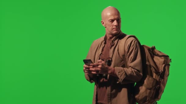 Turysta z plecakiem ubrany w khaki ubrania stojące przed zielonym ekranem chroma key, korzystający z mapy na telefonie komórkowym i rozglądający się podczas wędrówki - Materiał filmowy, wideo