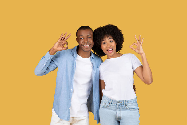 Gioiosa coppia afroamericana che fa bene segno con le mani, uomo in camicia blu e donna in camicia bianca, entrambi sorridenti felicemente contro uno sfondo giallo senape - Foto, immagini