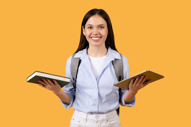 Estudante sorridente segurando livro em uma mão e tablet moderno na outra, fica contra fundo amarelo, simbolizando a interseção da educação clássica e digital - Foto, Imagem
