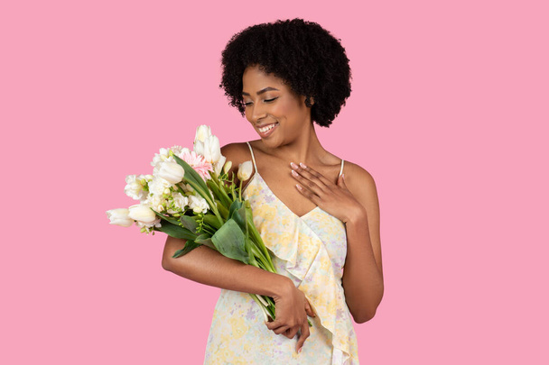 Content glad pretty millennial Afroamerykanka kobieta z naturalną fryzurę podziwiając piękny bukiet mieszanych kwiatów shes gospodarstwa, na miękkim różowym tle, studio - Zdjęcie, obraz