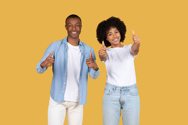 Щаслива афроамериканська пара дає великі пальці вгору, чоловік у блакитній сорочці і білі штани, а жінка в білій сорочці і джинсах, посміхаючись на жовтому тлі - Фото, зображення