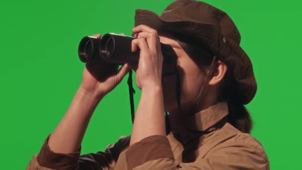 Молода жінка-туристка в хакі і капелюсі дивиться через бінокль під час походу в природу, стоячи на фоні ключа зеленої хроми - Кадри, відео