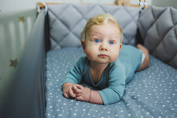 A 6-month-old child lies in a crib and looks at the camera. Gray bed linen for a baby bed - Photo, Image