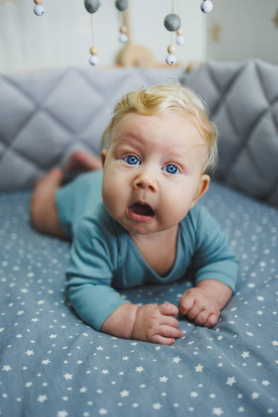 Ένα παιδί 6 μηνών βρίσκεται σε μια κούνια και κοιτάζει την κάμερα. Γκρι κλινοσκεπάσματα για παιδικό κρεβάτι - Φωτογραφία, εικόνα
