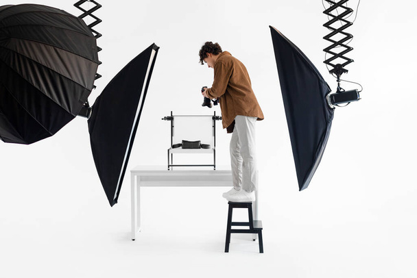 Focused male photographer stands on stool, meticulously adjusting his camera to capture the perfect studio shot, surrounded by professional lighting gear - Foto, Imagen