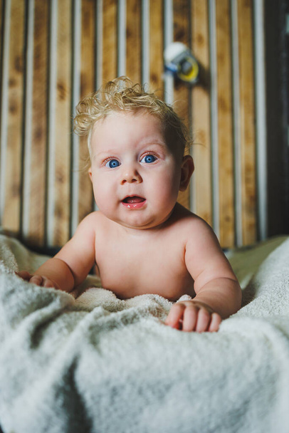 Το σγουρό νεογέννητο αγοράκι ξαπλώνει σε λευκή πετσέτα μετά το μπάνιο. Πορτρέτο ενός μωρού 6 μηνών με χαμόγελο στο πρόσωπό του. - Φωτογραφία, εικόνα