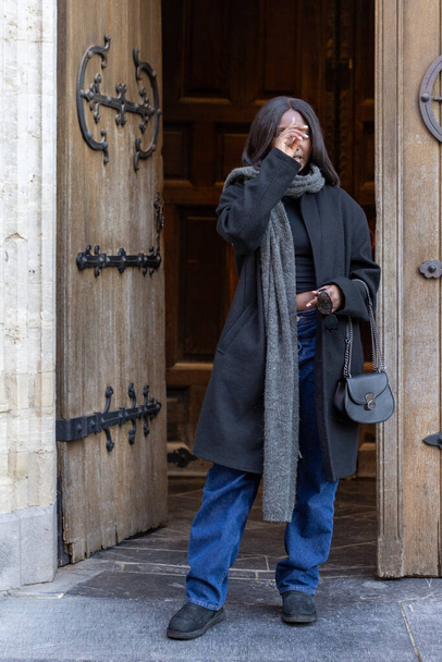 The image portrays a candid moment as a young woman, clad in a chic black coat and denim, steps out from the ancient doorway of a cathedral. With one hand delicately touching her face, she appears to - Photo, Image