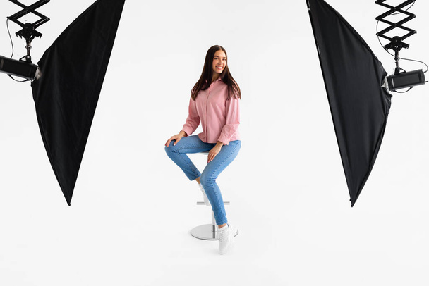 Młoda kobieta w miękkiej różowej bluzce i w niebieskich dżinsach uderza relaksującą pozę na stołku, otoczona profesjonalnym sprzętem oświetleniowym studio, na czystym białym tle - Zdjęcie, obraz