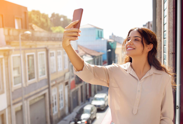 Beautiful young woman taking selfie with her smartphone on sunlit balcony, happy millennial female enjoying the vibrant city atmosphere, reflecting happy, carefree urban lifestyle, free space - Photo, Image