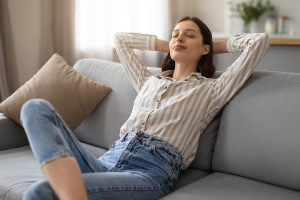 Rauhallinen nuori nainen nauttii rauhallinen hetki, makaa kädet pään takana pehmeällä sohvalla, johon sisältyy rentoutumista ja mukavuutta kotona, vapaa tila - Valokuva, kuva