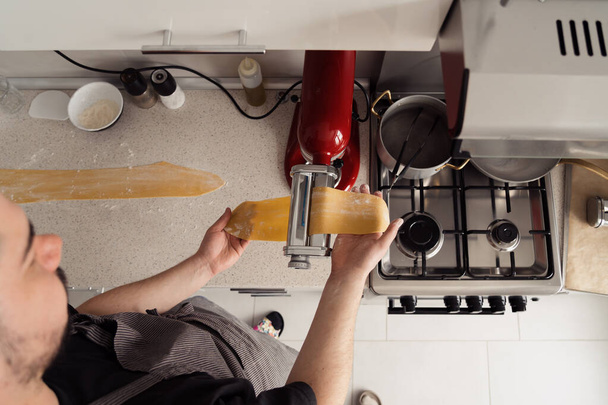 Κάτοψη του ατόμου που χρησιμοποιεί παρασκευαστή ζυμαρικών για να κυλήσει φρέσκια ζύμη σε μια οικιακή κουζίνα. - Φωτογραφία, εικόνα