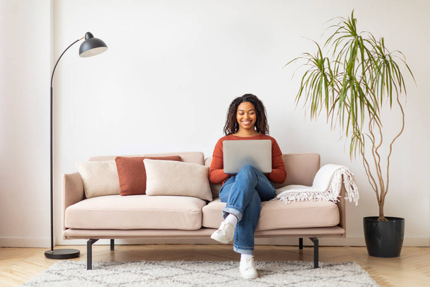 Carrera moderna. Mujer negra sonriente que trabaja con el ordenador portátil en casa, freelancer joven afroamericana feliz sentada en el sofá en el interior de la sala de estar y escribiendo en la computadora, espacio de copia - Foto, Imagen
