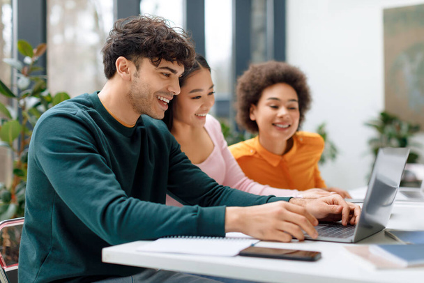 Három vidám diák különböző etnikai hátterű vesz részt együttműködési tanulmányi munkában laptoppal, buja beltéri növényekkel körülvéve. - Fotó, kép
