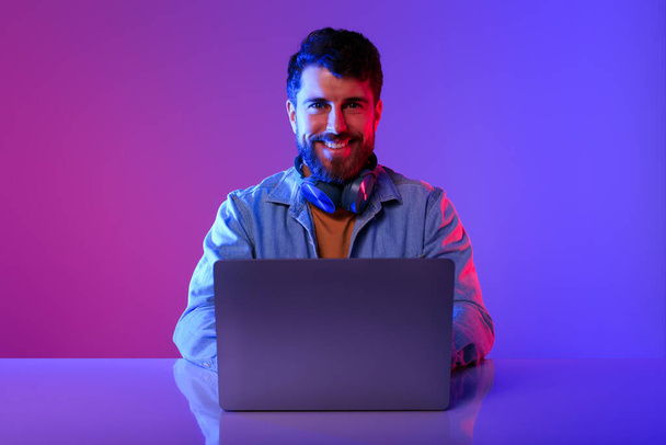 Online professioneel. Een blanke programmeur met een laptop aan zijn bureau, kijkend naar de camera, werkend op een computer in een paarse en blauwe neon studio. Internetcarrière, e-learningmogelijkheden - Foto, afbeelding