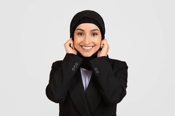 Сияющая молодая деловая женщина в профессиональной одежде, регулирующая свой хиджаб и смотрящая в камеру с радостным выражением лица, изолированная на светло-сером фоне - Фото, изображение