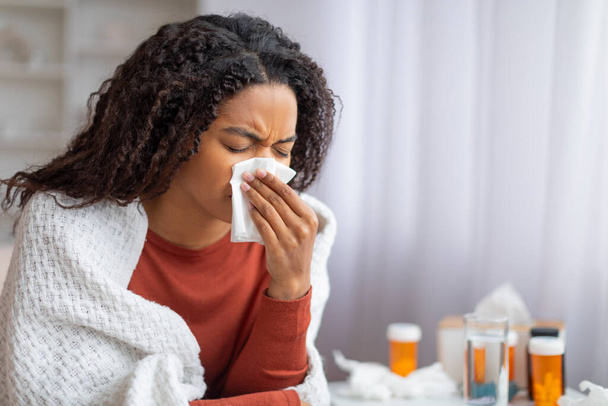 Niedobrze młoda czarna kobieta dmuchająca nos w chusteczkę w domu, chora afrykańska kobieta owinięta w koc, siedząca obok swoich leków, cierpiąca na przeziębienie i grypę, przestrzeń do kopiowania - Zdjęcie, obraz