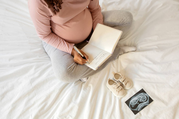 Mujer embarazada escribiendo pensamientos en su cuaderno en casa, sentada en la cama rodeada de pequeños zapatos y una imagen de ecografía, embarazada creando recuerdos durante su embarazo o haciendo una lista de verificación - Foto, imagen
