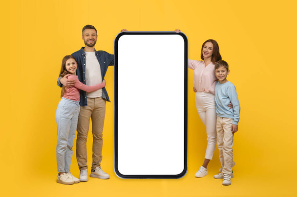 Szczęśliwa czteroosobowa rodzina stojąca blisko dużego smartfona z pustym ekranem, uśmiechniętymi młodymi rodzicami i ich dziećmi Reklama aplikacja mobilna, pozowanie razem na żółtym tle studio, kolaż, makieta - Zdjęcie, obraz