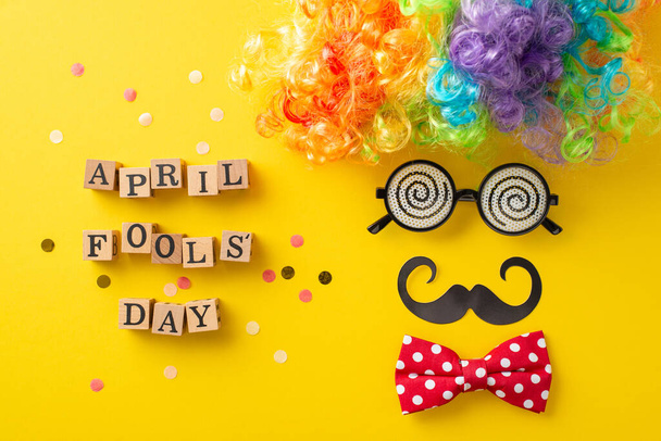 Prank top view set up, showcasing 'April Fool Day' spelt on cubes, along eximsical items like clown parig, specs, bow tie, mustaches, confetti, all arraated to mimic festive face on yellow backkdrop - Fotografie, Obrázek