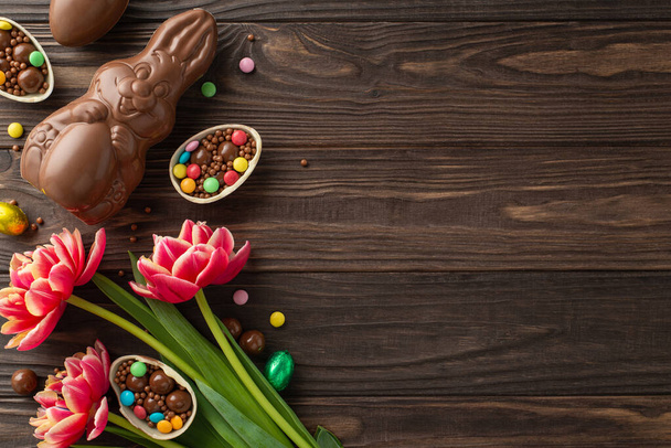 Charming Easter sweet treats concept. Overhead photo of chocolate eggs broken open with colorful candies spilling out, chocolate bunny, fresh tulips placed on wooden background, with space for text - Photo, Image